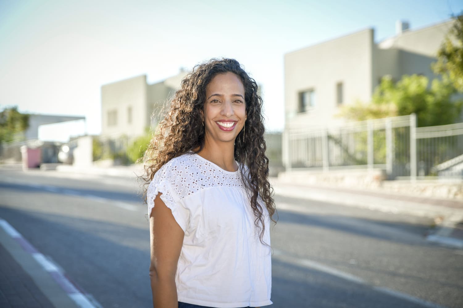 אישה מחייכת עם שיער מתולתל ארוך, לבושה חולצה לבנה, עומדת ברחוב שכונתי עם בתים ברקע