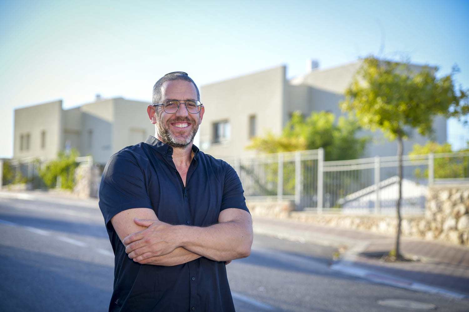 גבר מחייך עם משקפיים, זקן וכיפה, לבוש חולצה כהה, עומד בידיים שלובות ברחוב שכונתי עם בתים ברקע